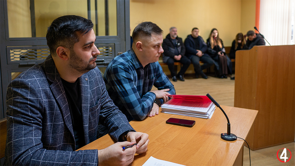 На фотографії зображено сцену у залі суду. На передньому плані за столом сидять двоє чоловіків. Ліворуч чоловік у темному піджаку в клітинку, праворуч – чоловік у картатій сорочці. На столі перед ними лежать стоси документів, червона папка, смартфон і мікрофон.  На задньому плані сидять інші люди, ймовірно, учасники або глядачі судового процесу. Приміщення має жовті стіни, дерев'яну підлогу та кафедру для виступів у правій частині кадру